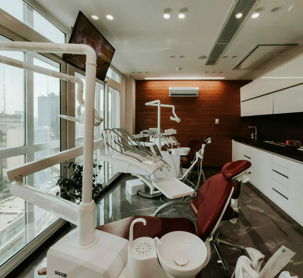 A modern dental clinic room with large windows offering natural light, featuring multiple dental chairs in burgundy and white, along with advanced dental equipment and tools. The space has a clean, minimalistic design with wood paneling on one wall and sleek white cabinets.