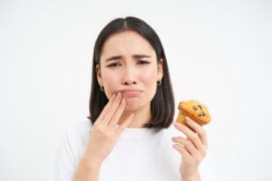 girl holding the outside of her right-side mouth because it hurts, while holding a muffin in her left hand