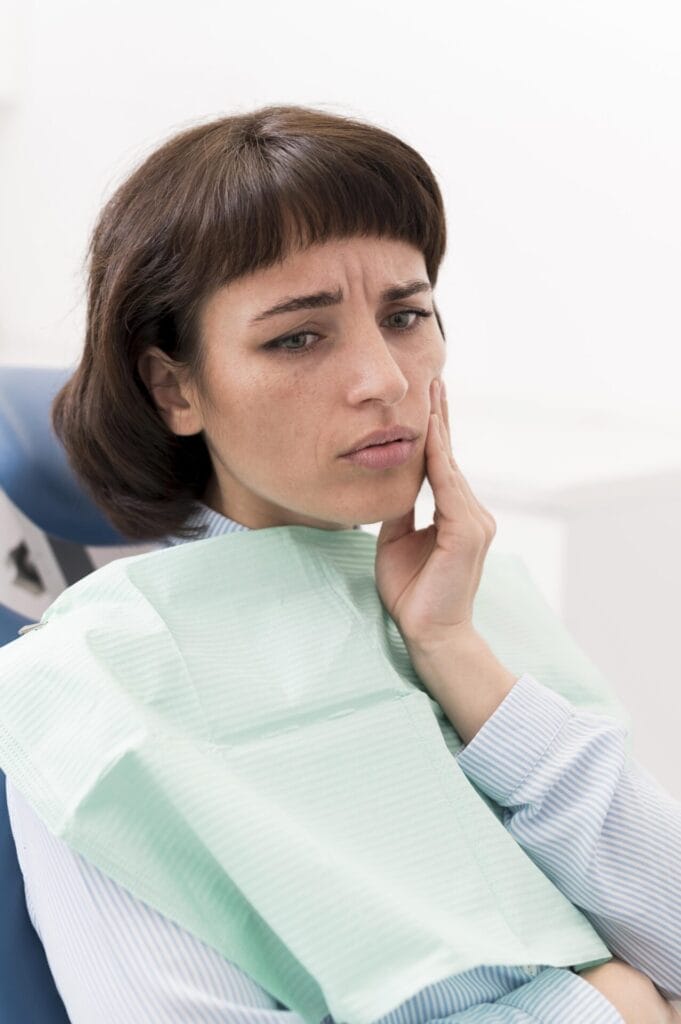 A woman sits in a dental chair, looking concerned and touching her cheek as if experiencing tooth pain or discomfort.