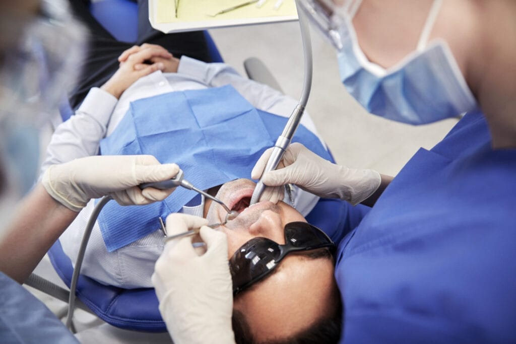 A patient wearing protective glasses lies in a dental chair while two dentists, wearing masks and gloves, work on his teeth using dental instruments.