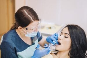 Dental professional wearing protective gear and face shield administering local anesthesia injection to a patient before an oral surgery procedure in a clinical setting.