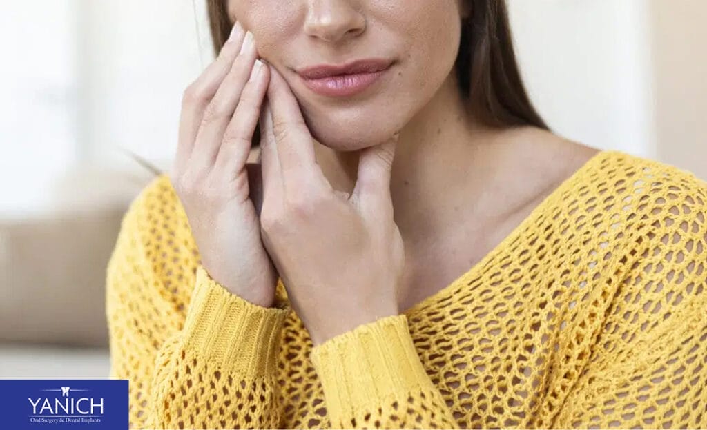Female patient in a yellow sweater touching her cheek, showing visible discomfort and dental pain that may require oral surgery or advanced treatment.
